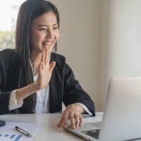 Happy young asian business woman waving hands to greeting partner during making video conference with her team.